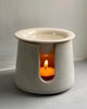A ceramic burner with a white finish, featuring a lit candle inside, placed on a flat surface.