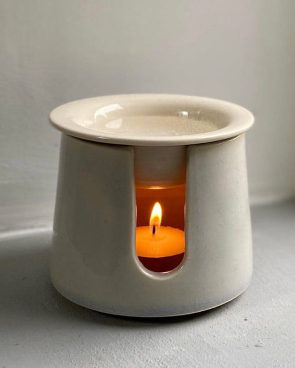 A ceramic burner with a white finish, featuring a lit candle inside, placed on a flat surface.