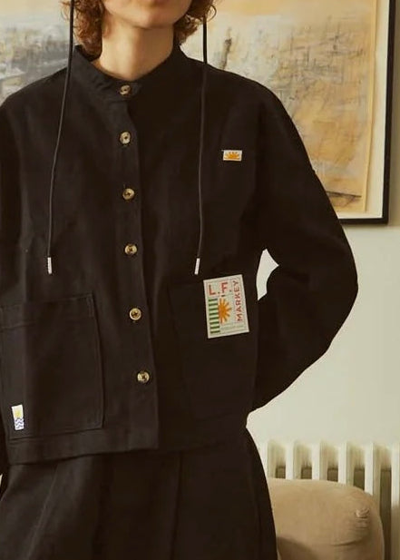 A model stands in a living room wearing a black cropped button-up jacket with drawstrings and gold-tone buttons, paired with black wide-leg pants and a black cap. The jacket features a small yellow logo patch on the chest and a larger white brand tag on the pocket.