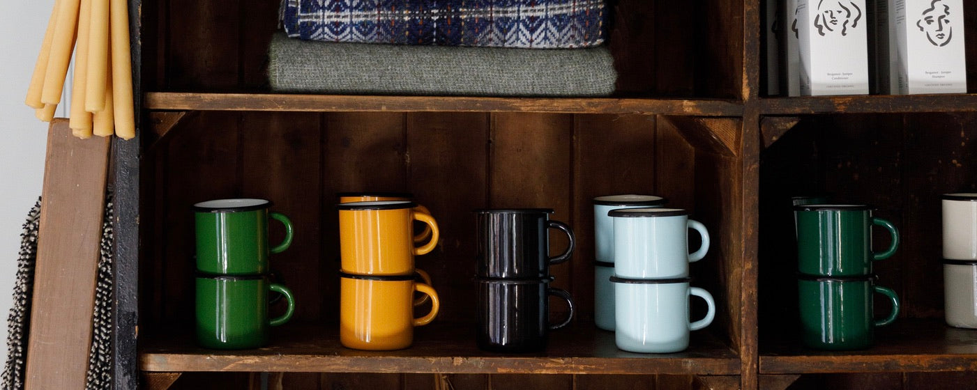 A collection of stacked ceramic mugs in various colors including green, orange, black, light blue, and dark green, displayed on wooden shelves alongside folded textiles and packaged goods.